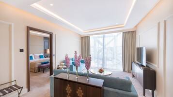 A modern hotel room with a sofa, a TV, a dining table, and a large window with curtains.