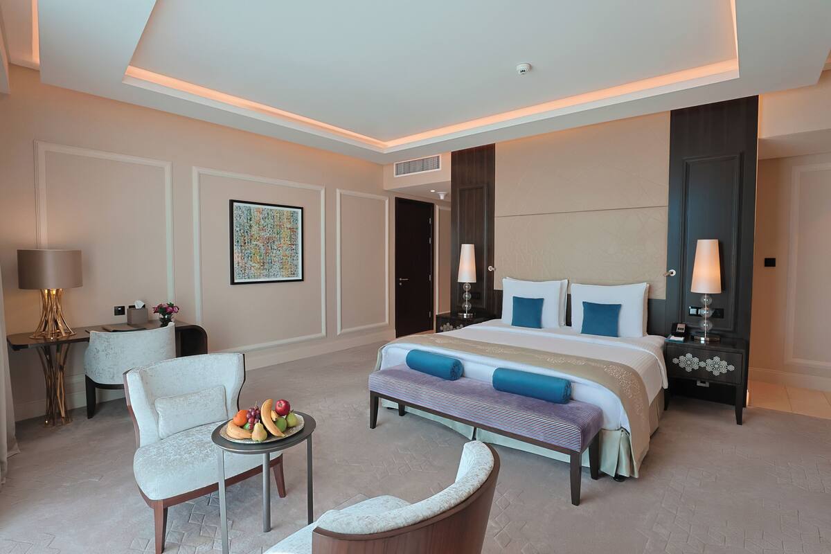A modern hotel room with a large bed, bedside tables, a seating area with a table, and a decorative wall piece.