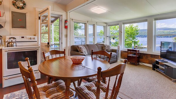 A spacious living area with a large dining table, comfortable seating, a kitchen with a stove, and a view of a lake through large windows.