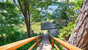 A wooden staircase leading to a building with a sloped roof, surrounded by lush greenery and a view of a lake.