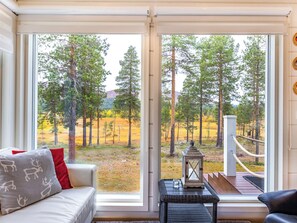 Ein Zimmer mit großem Fenster, einer Couch mit gemusterten Kissen, einem kleinen Tisch mit einer Laterne und Blick auf eine bewaldete Landschaft.