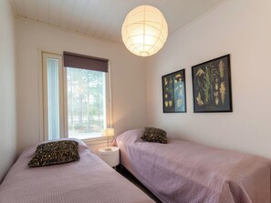 A bedroom with two beds, a window with blinds, a bedside lamp, and two framed botanical prints on the wall.