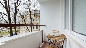 Un balcón con mesa y sillas de madera, una ventana con persianas y vistas a edificios y árboles.