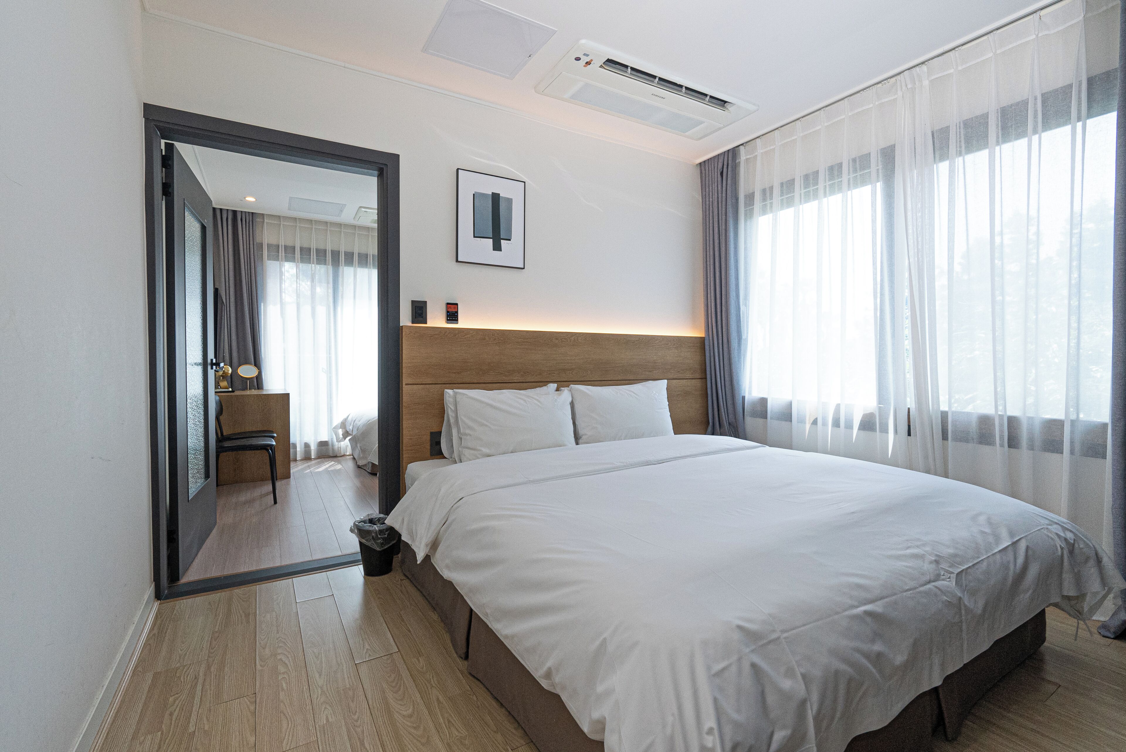 A hotel room with a large bed, a wooden headboard, a desk with a chair, and a large window with sheer curtains.