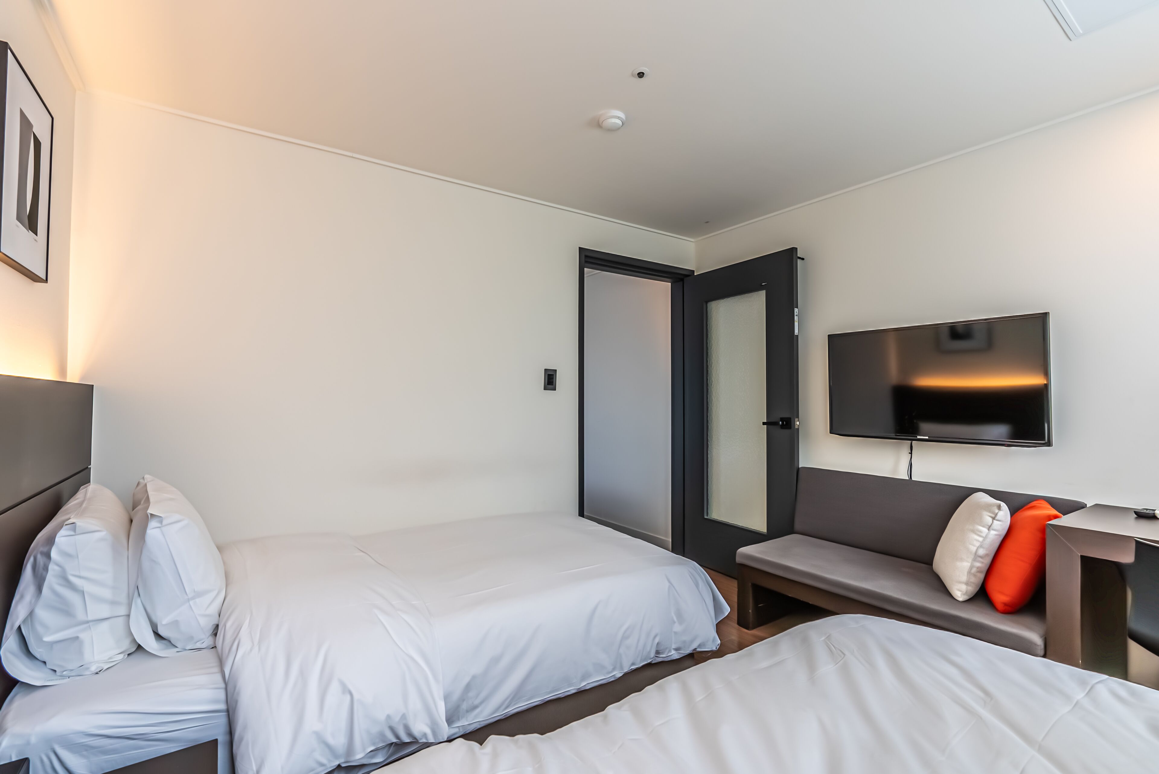A modern hotel room with two single beds, a sofa, a flat-screen TV, and a door leading to another room.