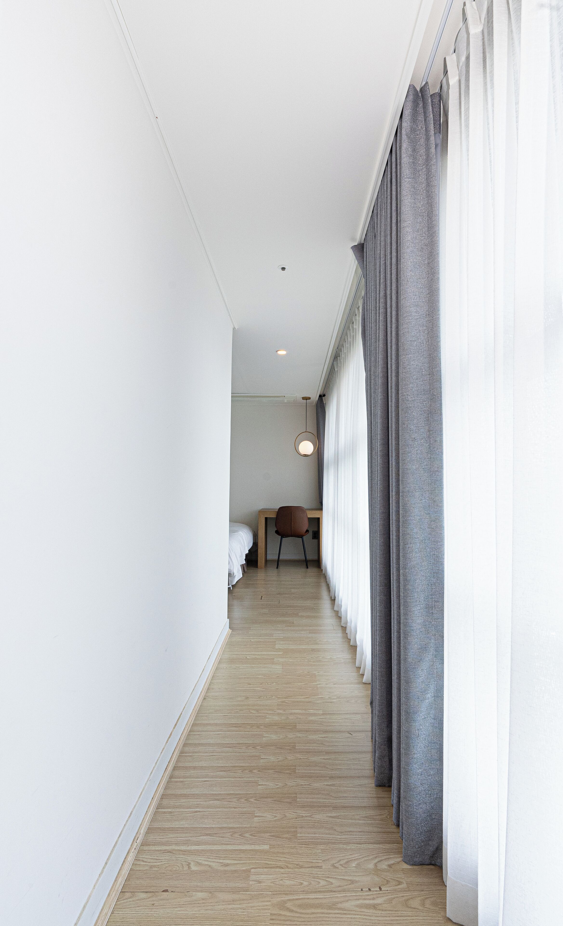 A narrow hotel corridor with a wooden floor, a bed, a chair, and a hanging light fixture.