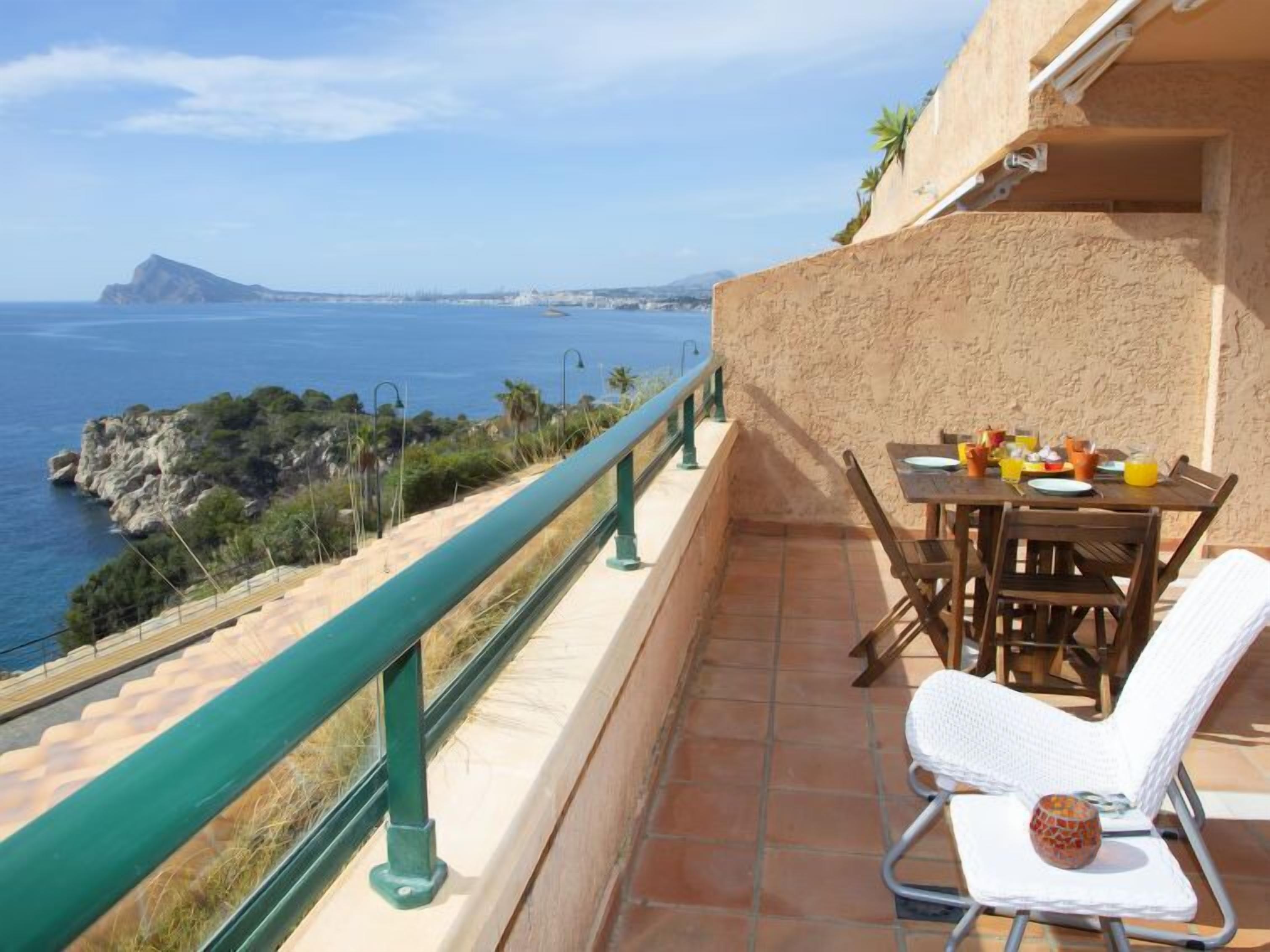 A balcony with a table set for breakfast, overlooking a coastal landscape with a view of the sea and a distant island.