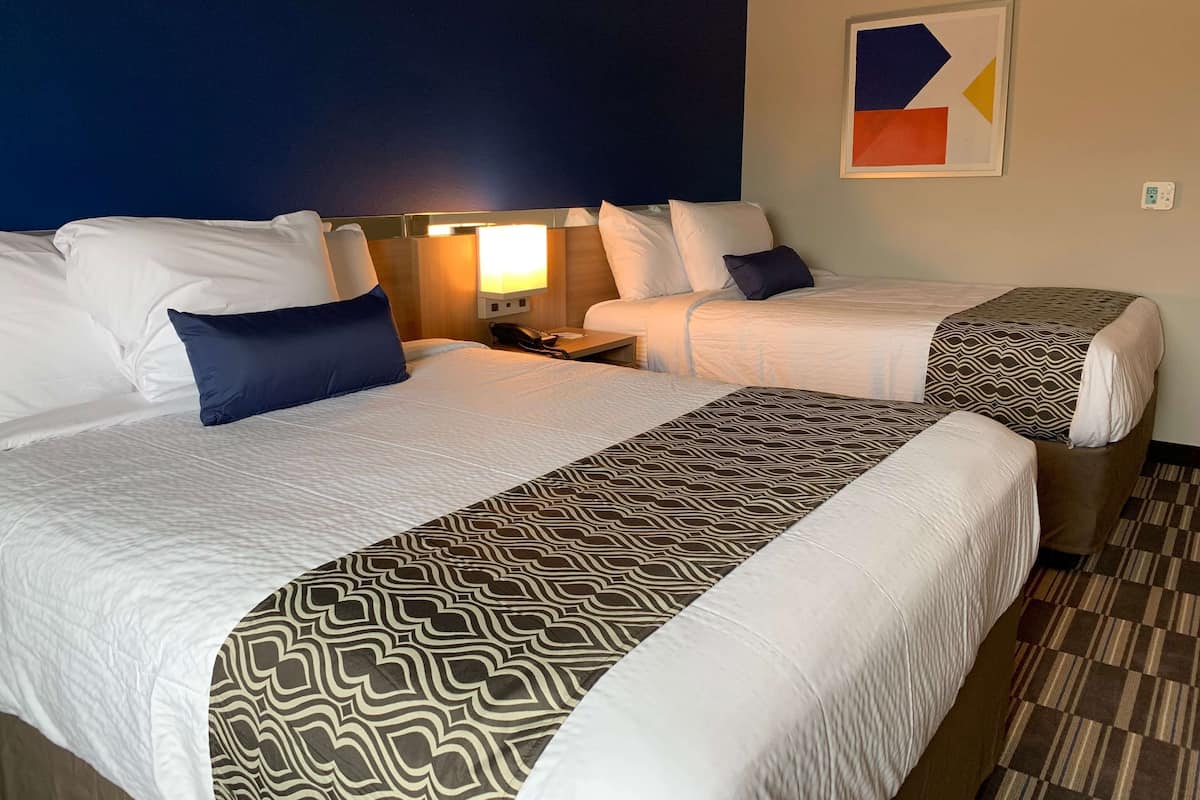 A hotel room with two beds, each with a patterned bedspread, a bedside table with a lamp, and a wall-mounted abstract art piece.