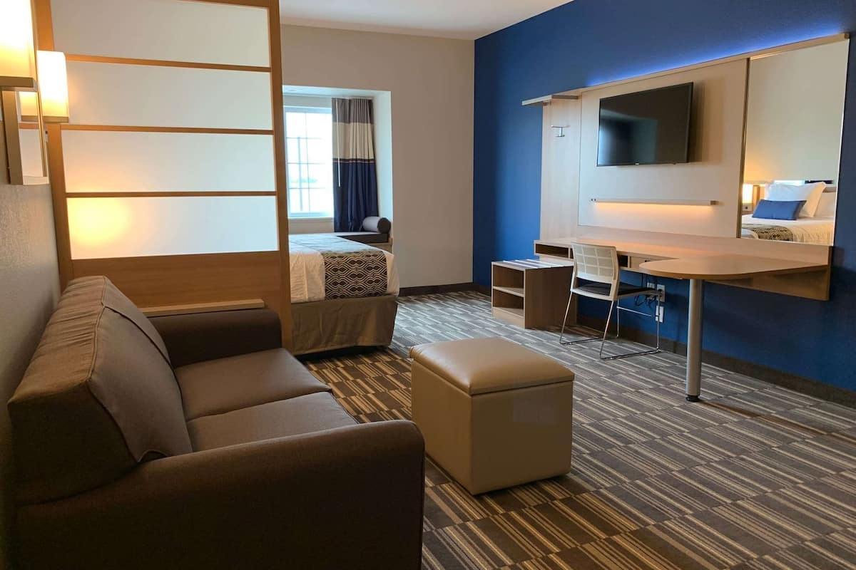 A hotel room with a sofa, ottoman, desk, chair, TV, and a bed with a window view.