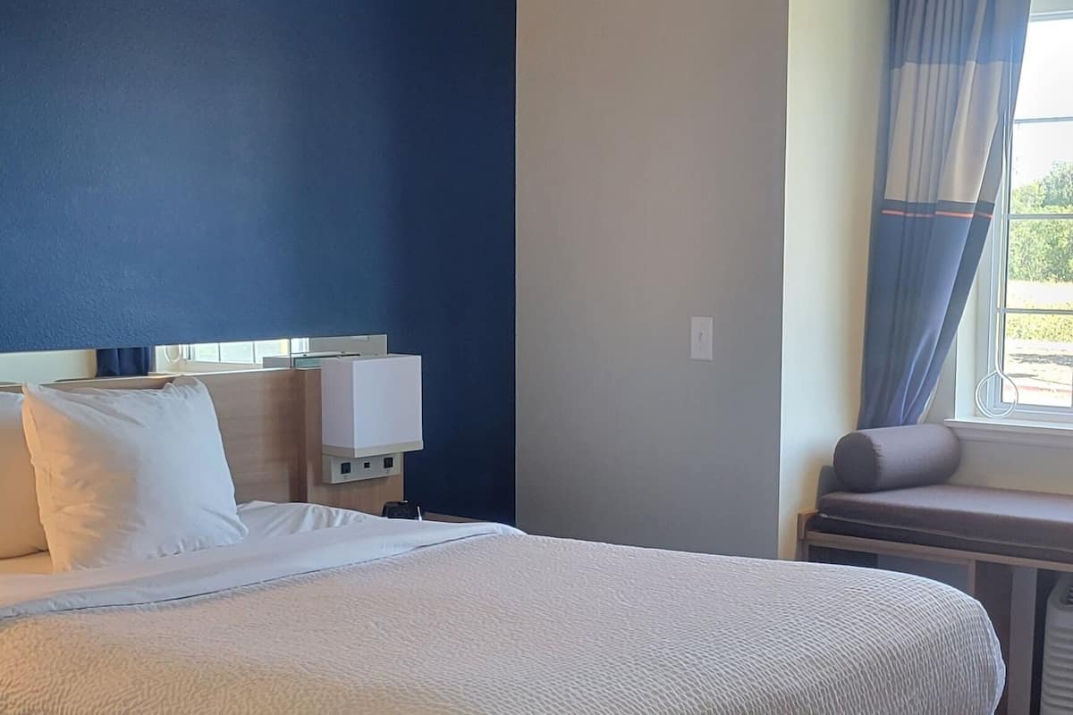 A neatly made bed with a white comforter and a brown skirt, a bedside table with a lamp, and a window with curtains.