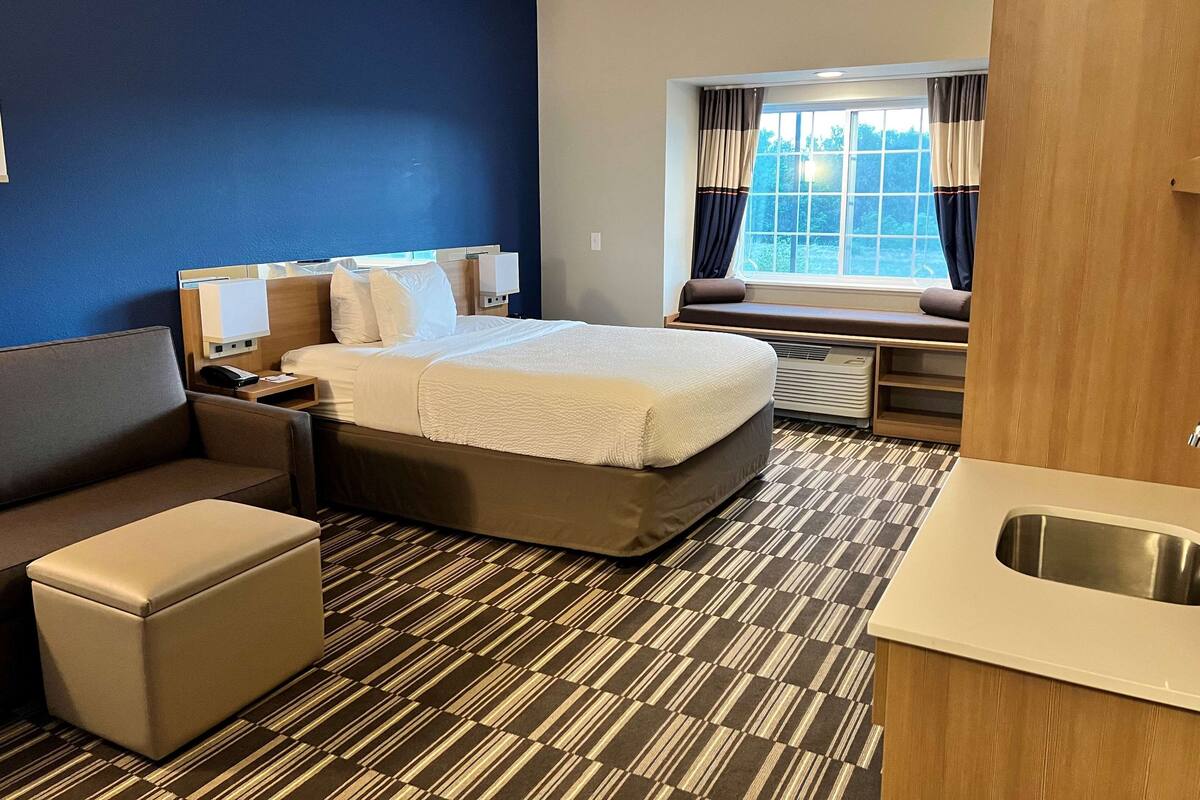 A hotel room with a bed, a sofa, a small table, a nightstand, a window with curtains, and a kitchenette area.