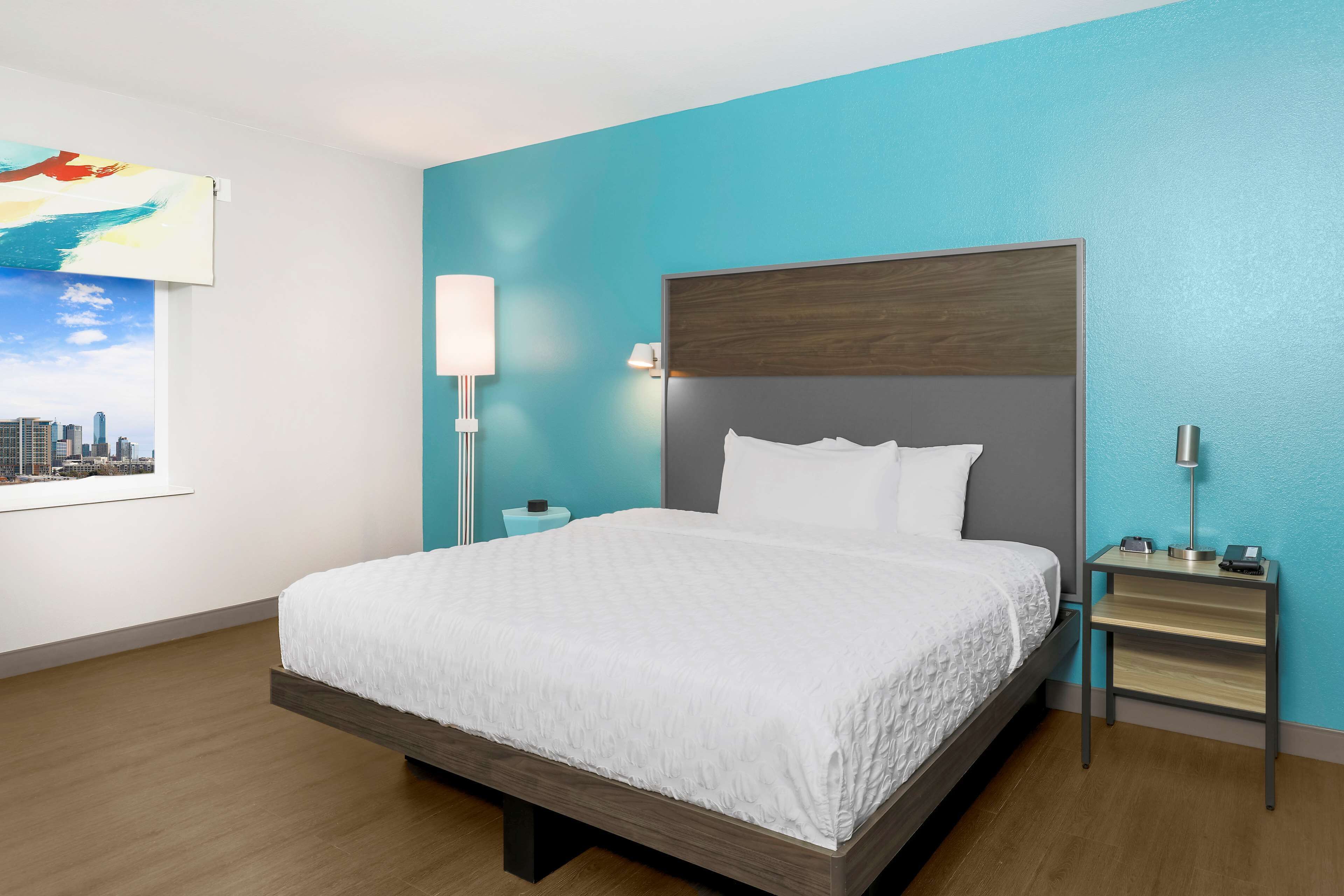 A modern hotel room with a large bed, a nightstand, a wall-mounted lamp, and a cityscape painting.