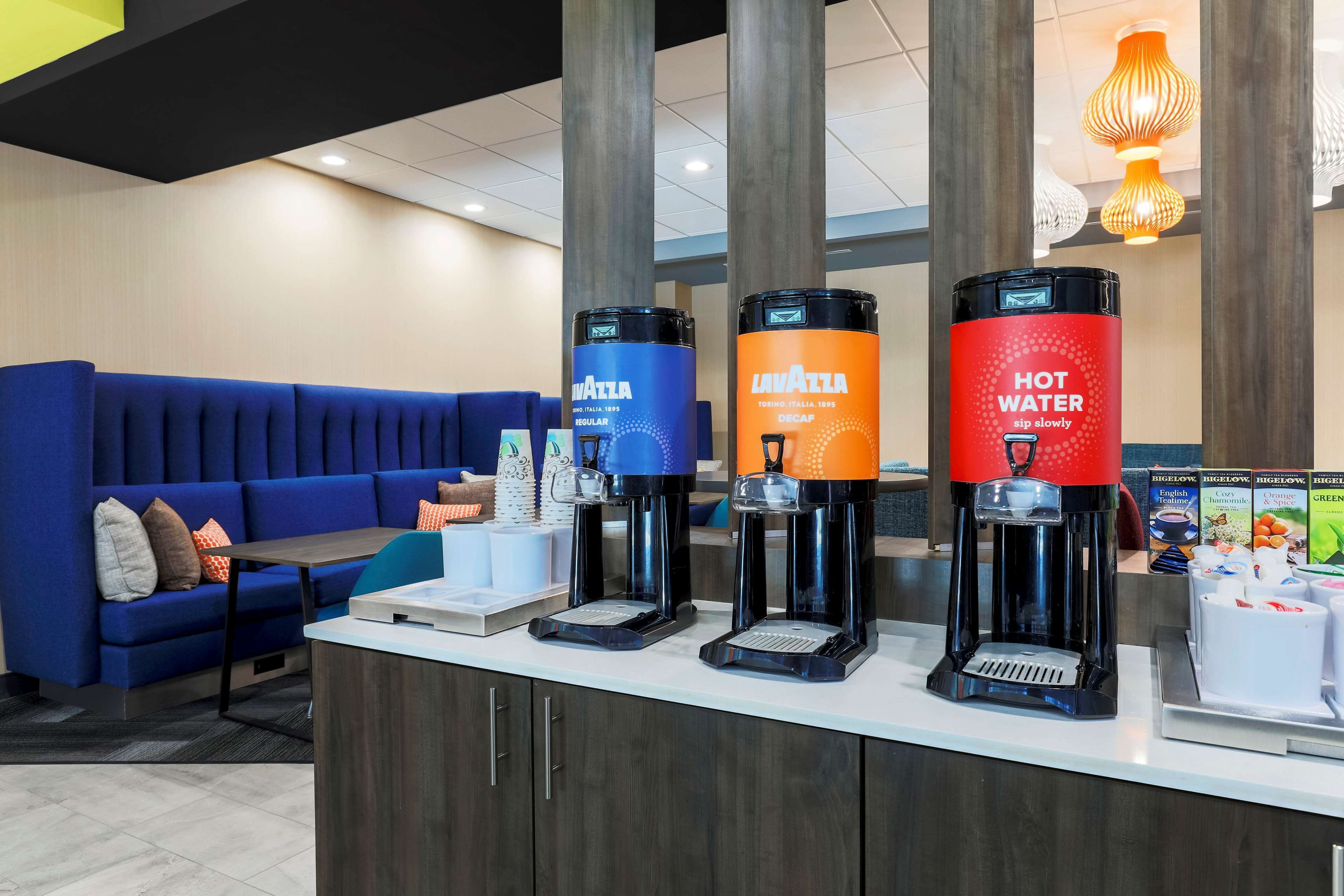 A hotel reception area with a blue sofa, a coffee station with three dispensers, and a counter with a coffee machine.