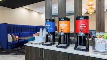 A hotel reception area with a blue sofa, a coffee station with three dispensers, and a counter with a coffee machine.