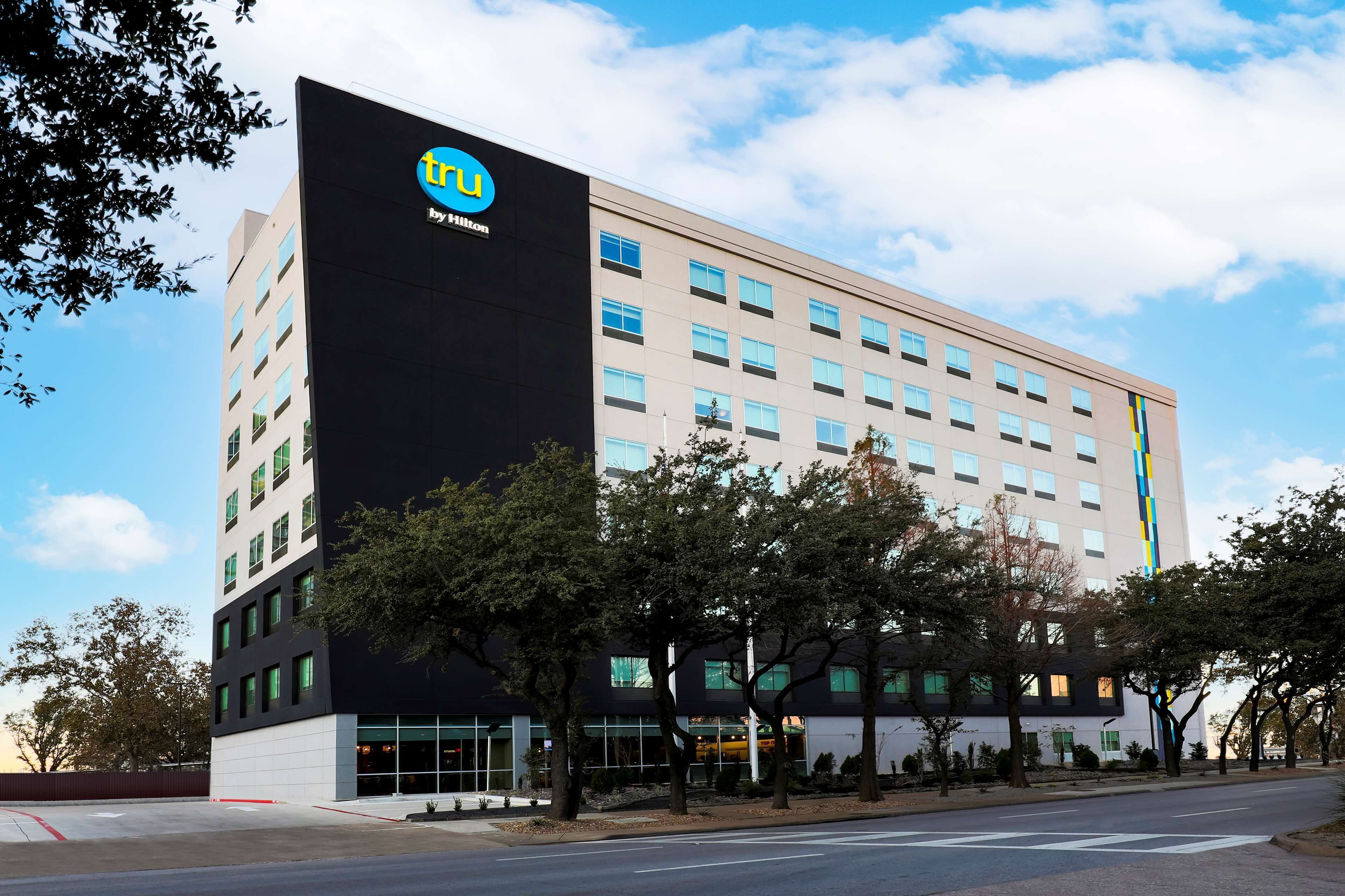 Un moderno edificio de hotel con un llamativo letrero de 'TRU by Hilton', varios pisos y un estacionamiento al frente.