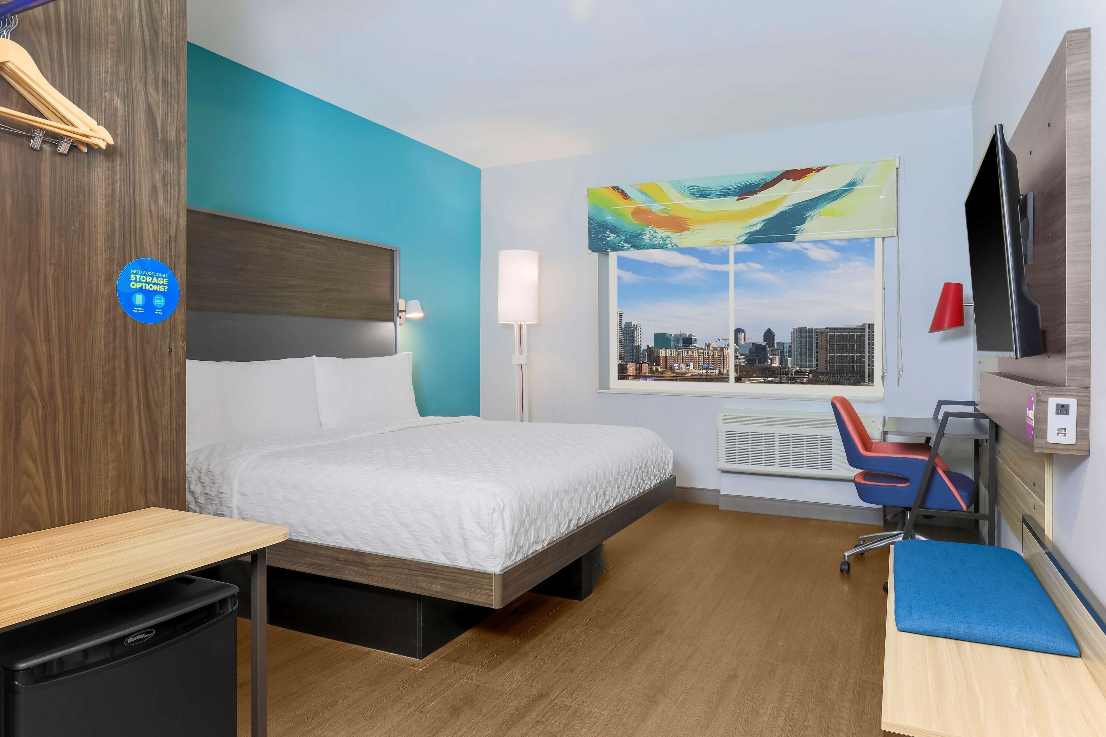 A hotel room with a bed, a desk, a chair, a TV, a window with a cityscape view, and a painting on the wall.