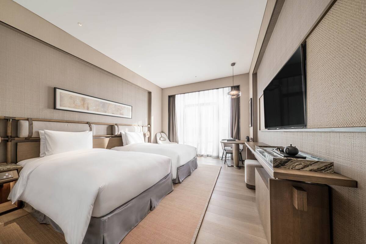 A modern hotel room with two beds, a desk, a chair, a large window, and a flat-screen TV.