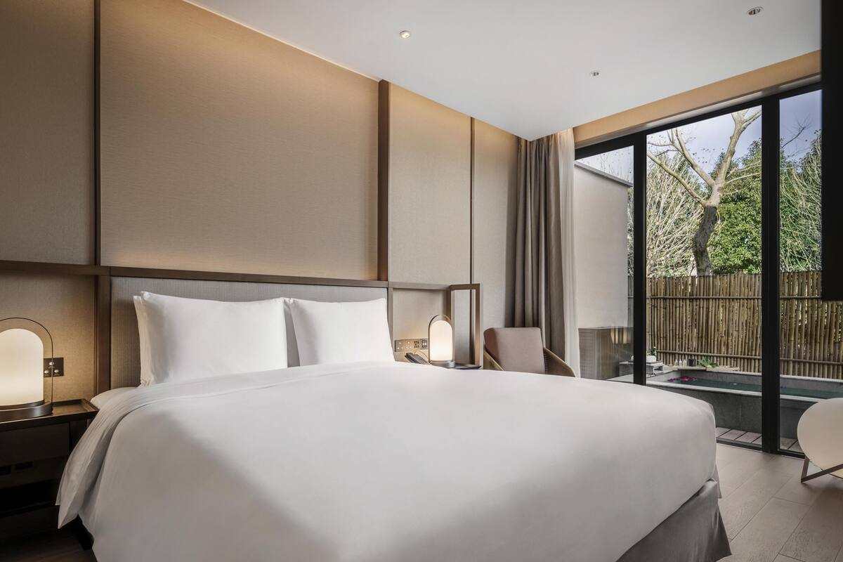 A modern hotel room with a large bed, a bedside table with a lamp, a chair, and a view of the outdoors through a sliding glass door.