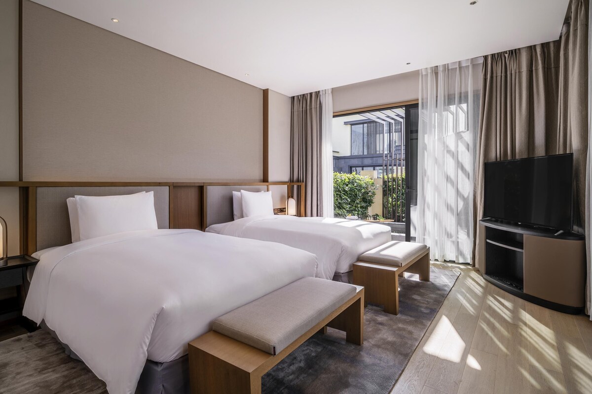 A modern hotel room with two beds, a bench, a TV, and a view of greenery outside.