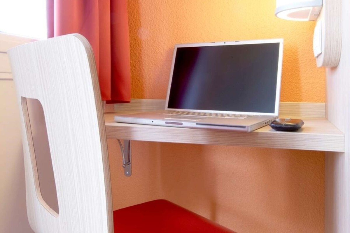 Ein Laptop auf einem Schreibtisch in einem Zimmer mit einem roten Stuhl und einem Fenster mit Vorhängen.