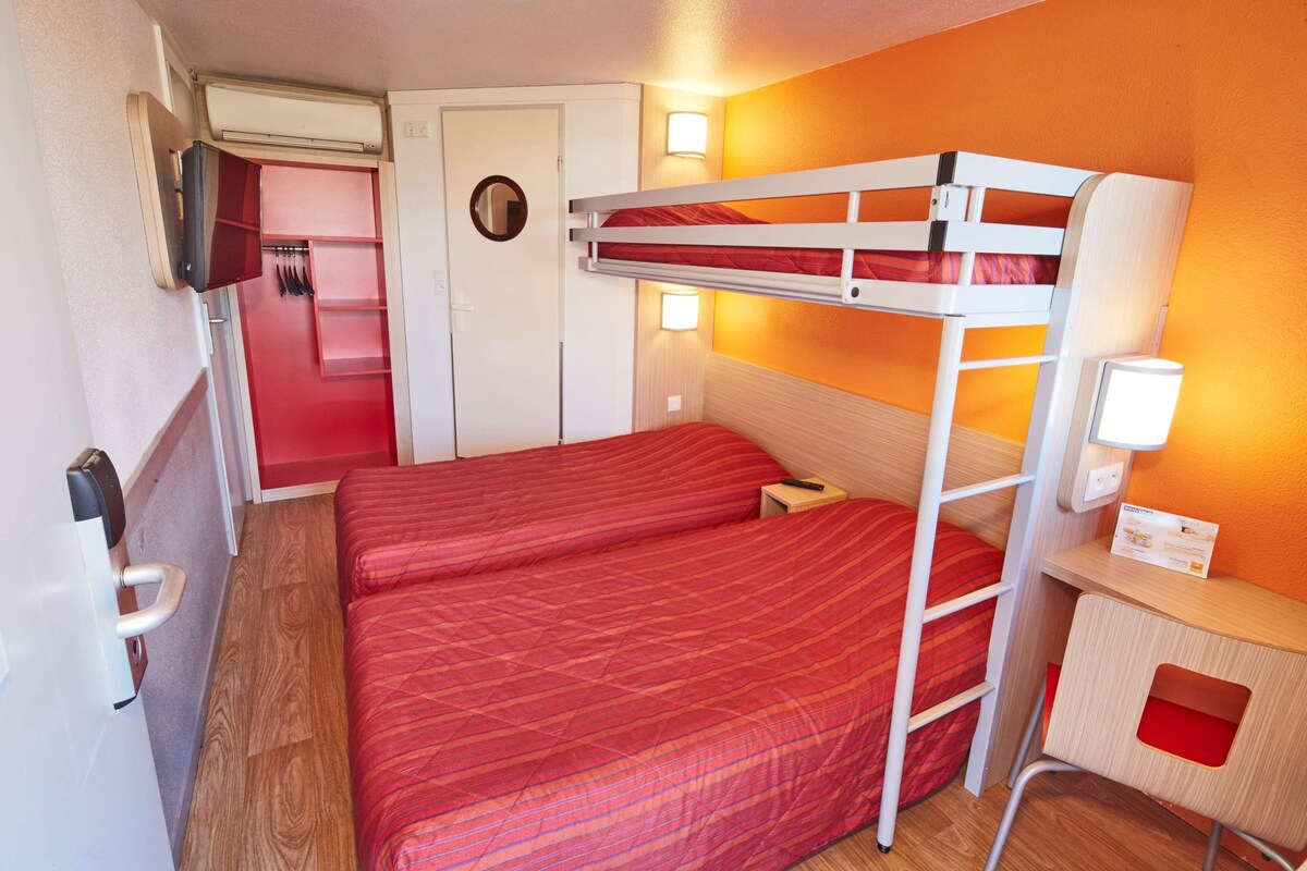 Ein Schlafzimmer mit Etagenbetten, einer roten Bettdecke, einem kleinen Schreibtisch mit Lampe und einer Tür mit Spiegel.