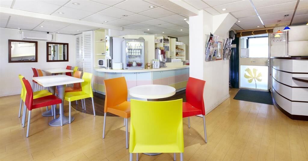 Un espacio interior luminoso con una mesa redonda central rodeada de sillas coloridas, una encimera con un refrigerador y una puerta con un diseño de hojas.