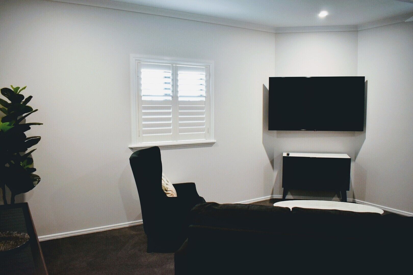 Una habitación con televisión de pantalla plana montada en la pared, una ventana con persianas, una silla negra y una pequeña mesa.