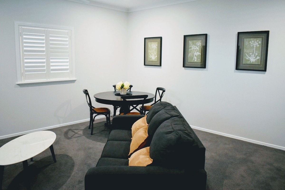 A room with a black sectional sofa, a round dining table with chairs, and three framed botanical prints on the wall.