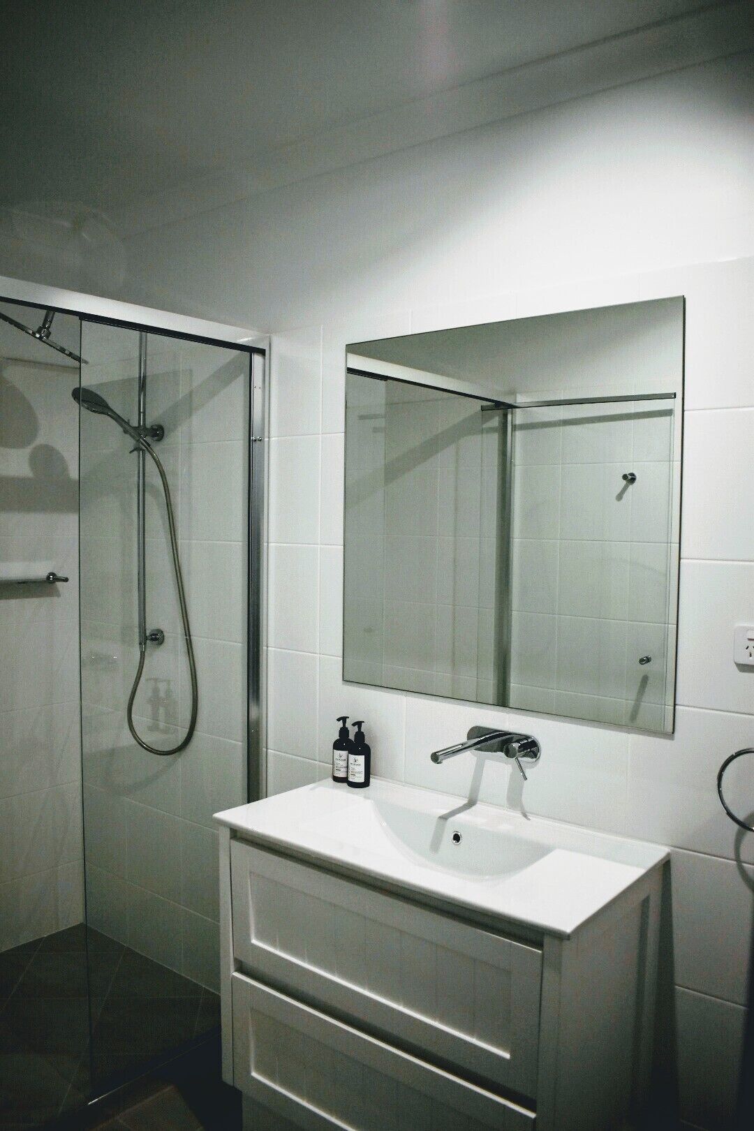 A modern bathroom with a glass-enclosed shower, a white sink with a mirror above it, and two bottles of shampoo on the sink.