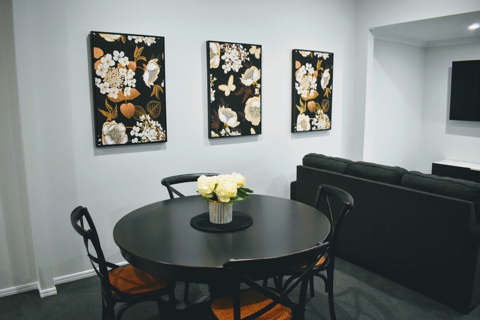 Un coin repas avec une table ronde, des chaises et un vase de fleurs. Trois tableaux sont accrochés au mur au-dessus de la table.