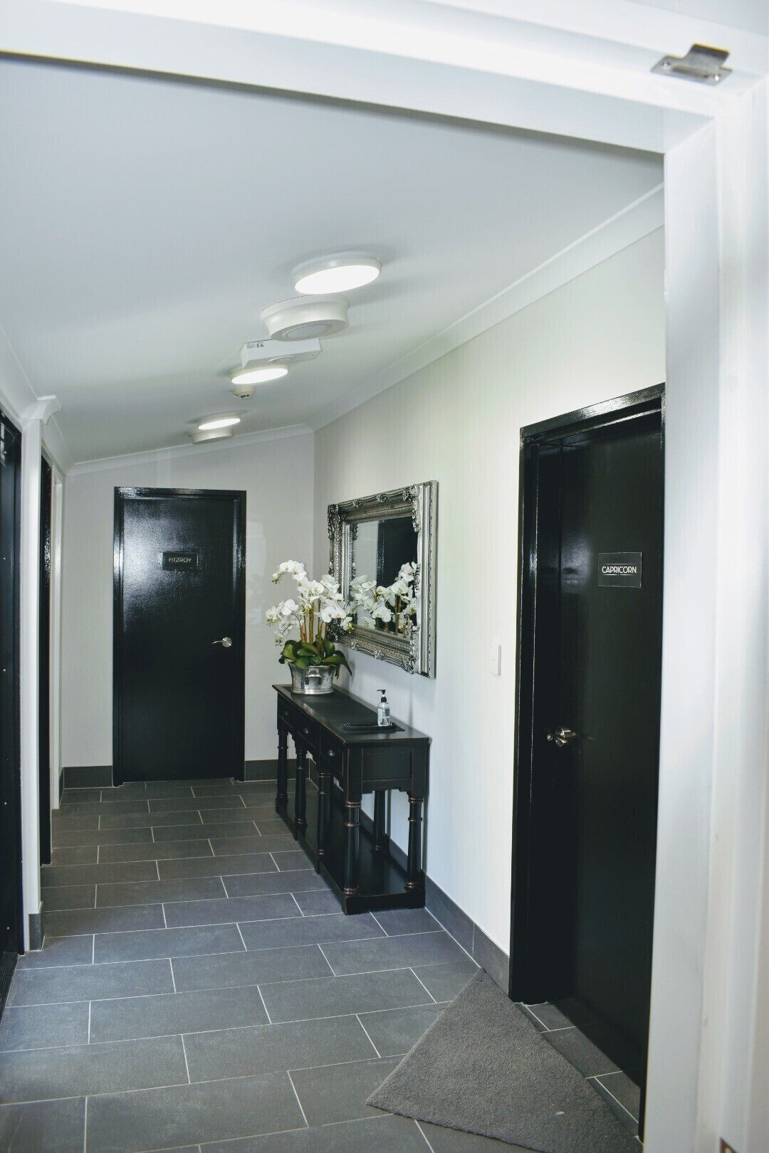 A hallway with black doors, a mirror, and a vase of white flowers on a console table.