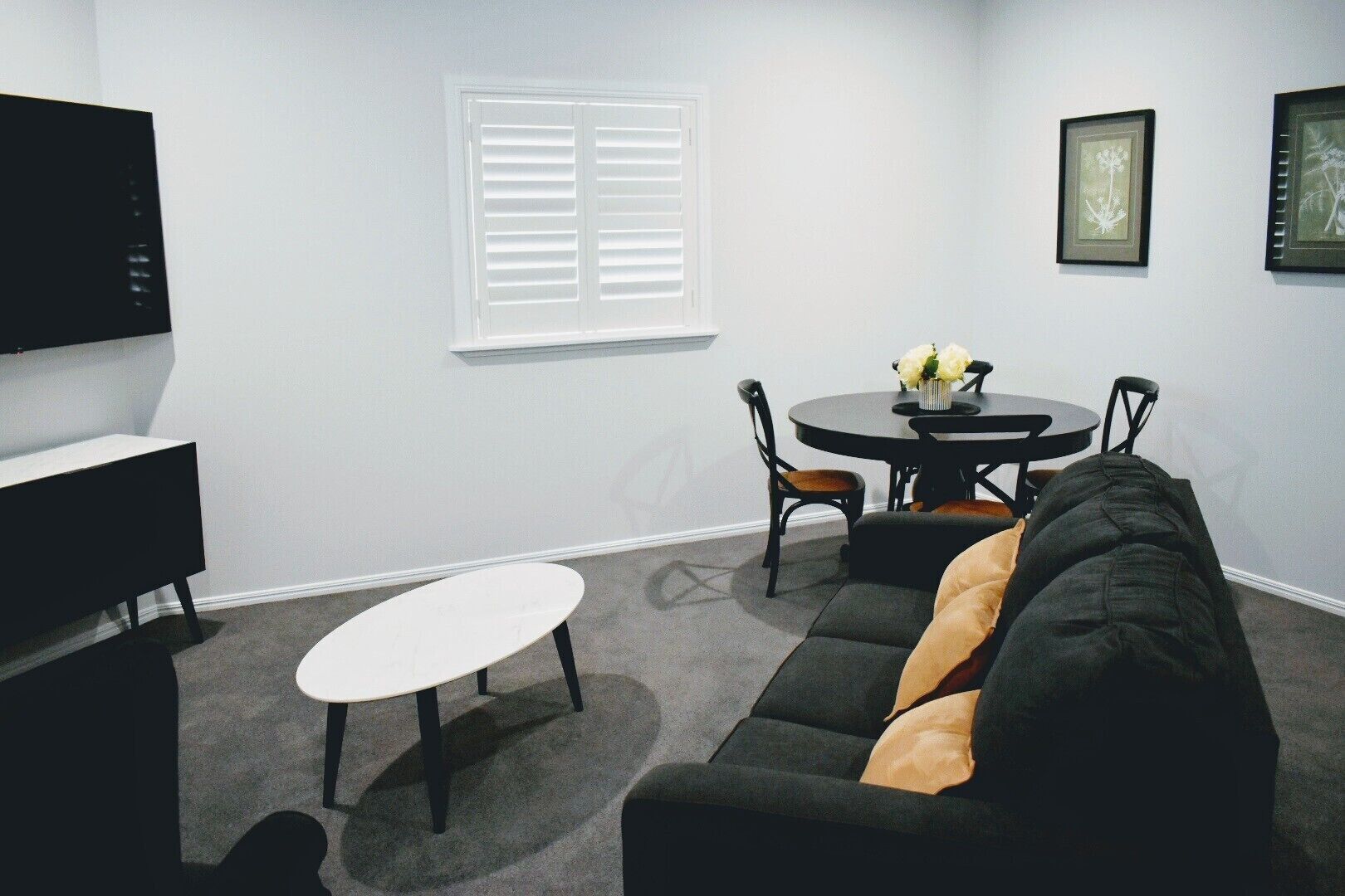 A modern living room with a black sofa, a round dining table with chairs, a white TV stand, and a window with blinds.