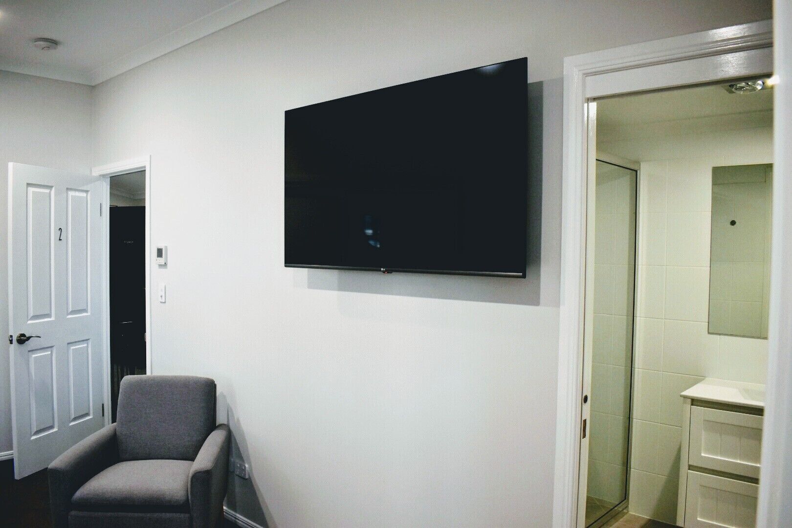 Une pièce dotée d’un téléviseur à écran plat fixé au mur, d’un fauteuil gris et d’une porte blanche donnant accès à une salle de bain.