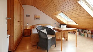 A cozy attic bedroom with a sloped ceiling, wooden walls, a bed, a wicker chair, a small table, and a bookshelf.