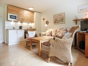 A compact living space with a kitchenette, a sofa, a wicker chair, and a television. - Urlauberdorf 25b, Deutschland (Ostseebad Boltenhagen)