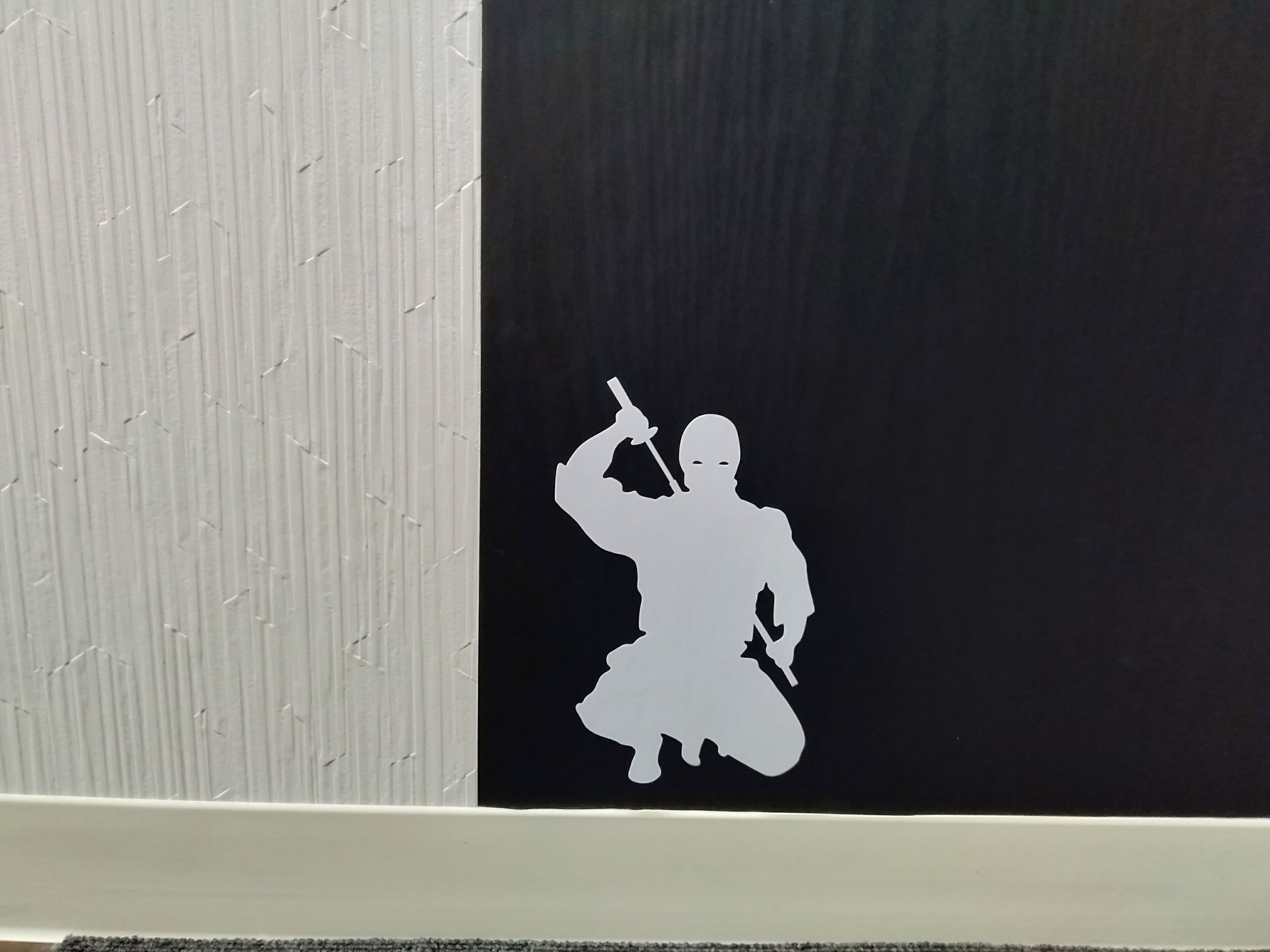 Een muur met een wit reliëf aan de linkerkant en een zwart gedeelte met een witte silhouet van een persoon die een zwaard vasthoudt aan de rechterkant.