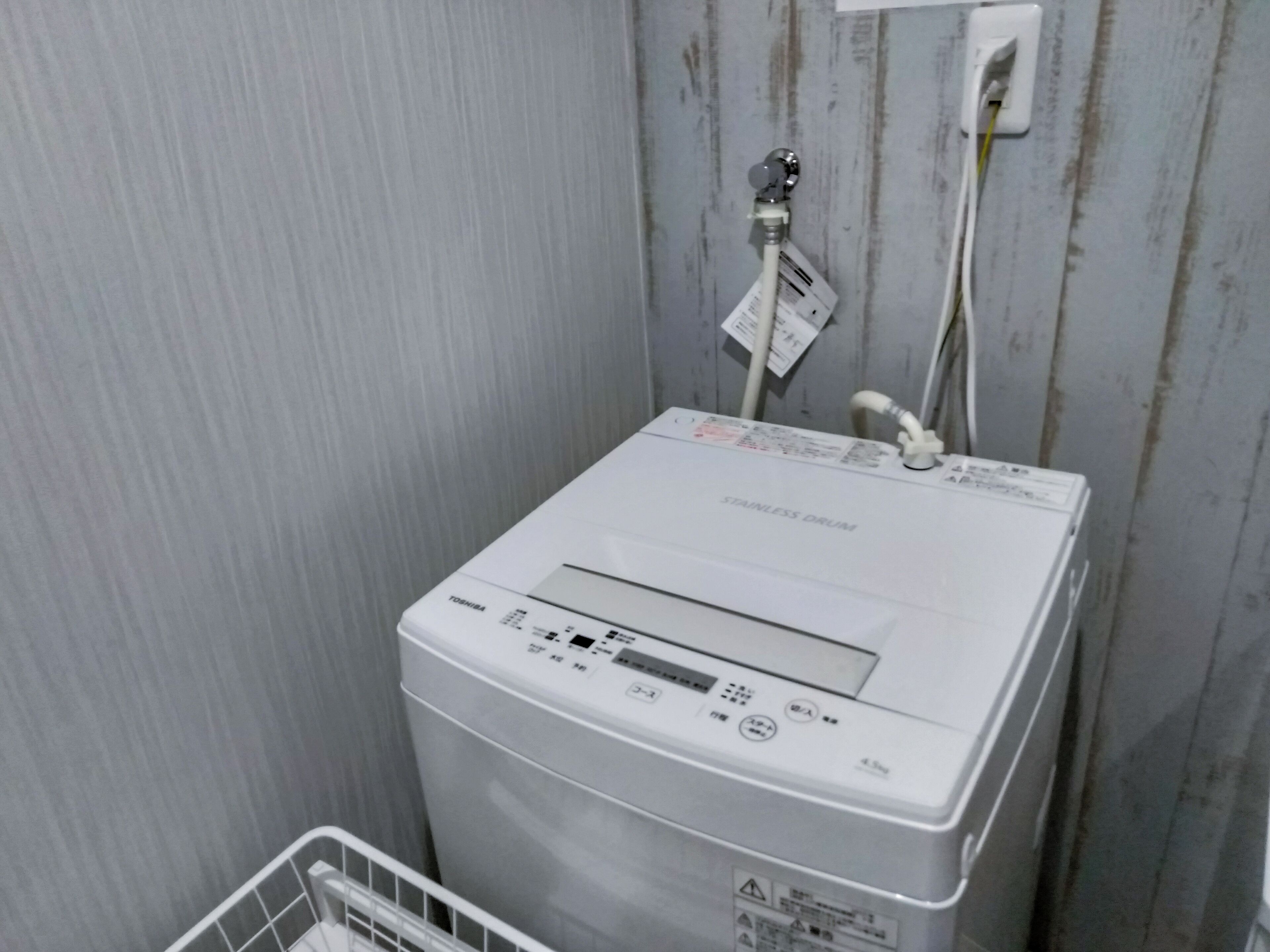 A white, stainless steel drum water heater with a digital display and control panel, mounted on a wall with visible pipes and a power outlet.
