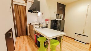 A compact kitchen with a white countertop, a microwave, a refrigerator, and a sink. There are two green stools and a small trash bin. - Sakura Villa Osaka (Osaka)