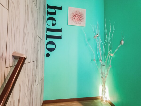 Una escalera con barandilla, una pared verde azulado, una obra de arte floral enmarco y un jarrón decorativo con ramas y adornos esféricos.