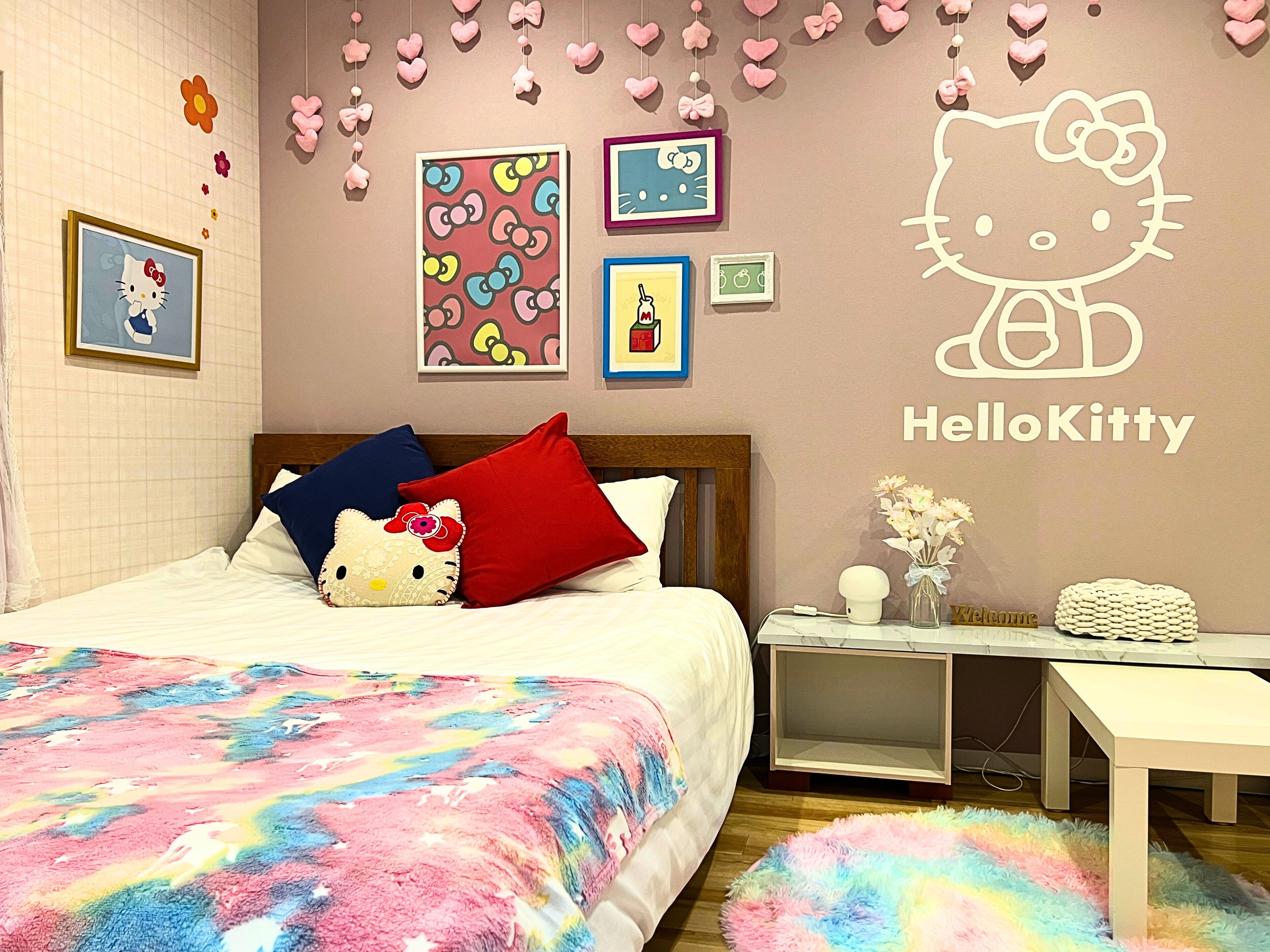 Une chambre à coucher avec un lit, des oreillers, une table de chevet, une commode et un mur décoré avec des motifs de Hello Kitty.