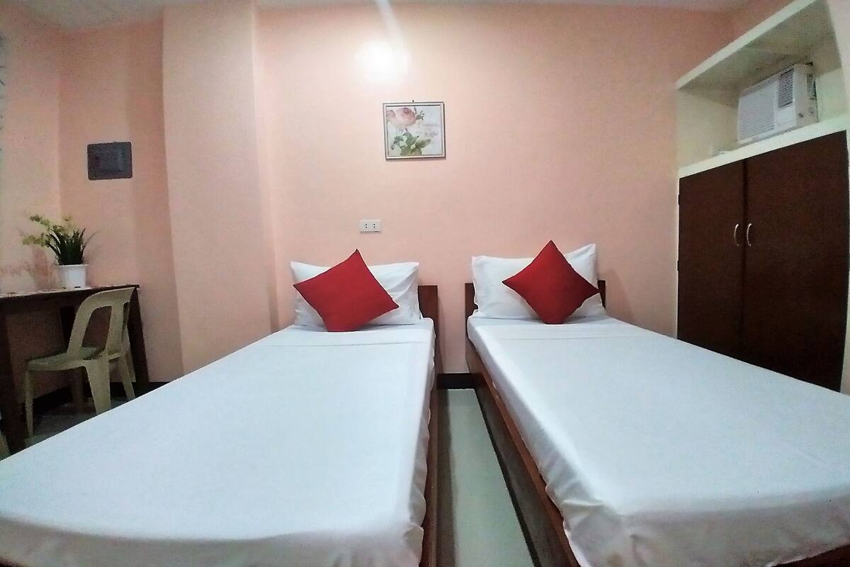A room with two single beds, each with a red pillow, a desk with a chair, and a shelf with an air conditioner.