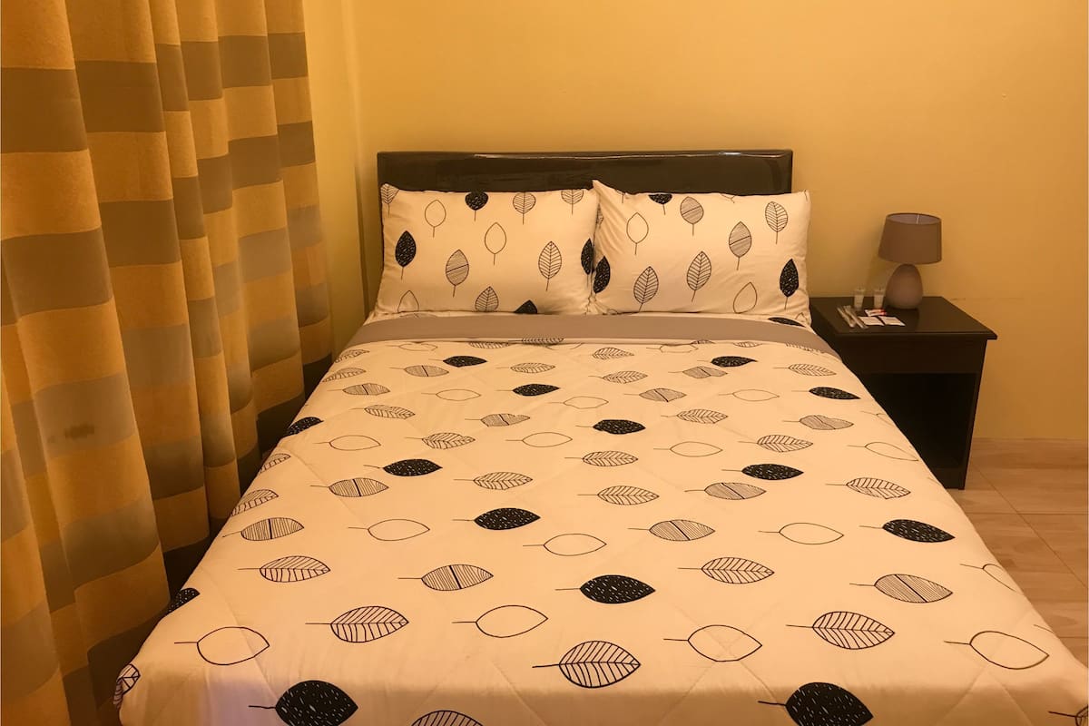 Una cama bien hecha con una colcha estampada de hojas, una mesita de noche con lámpara y una cortina ligeramente entreabierta.