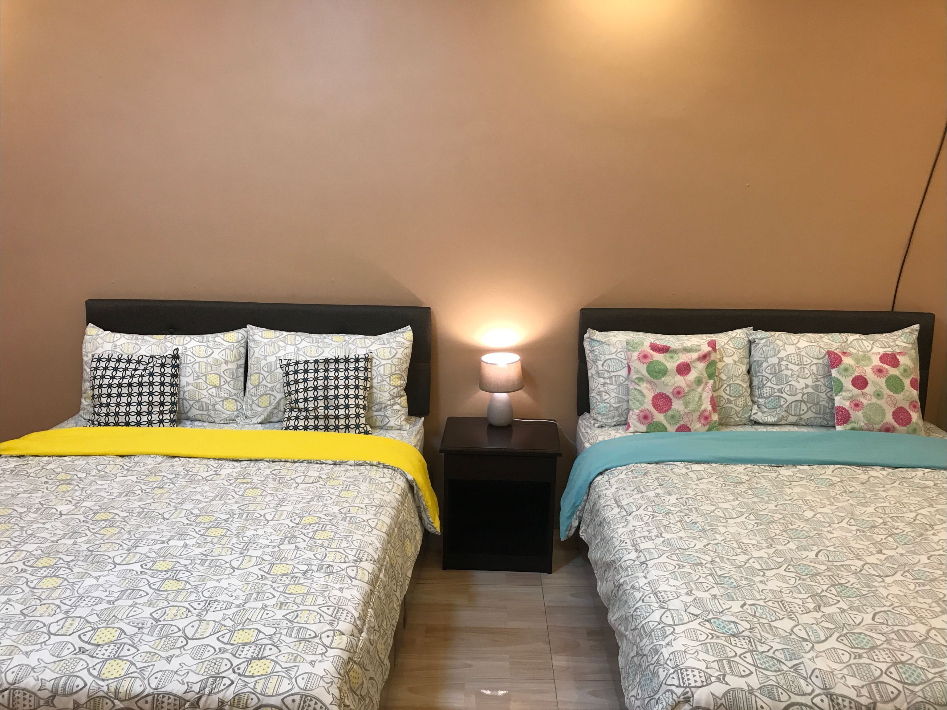Quarto com duas camas, cada uma com roupa de cama e almofadas estampadas, mesa de cabeceira com abajur e chão de madeira.