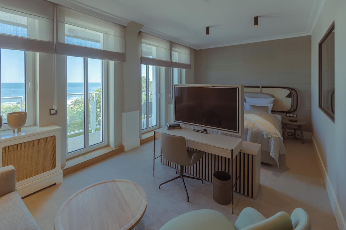 Ein modernes Hotelzimmer mit großem Fernseher, einem Bett mit weißem Kopfteil, einem Schreibtisch und Meerblick durch große Fenster.