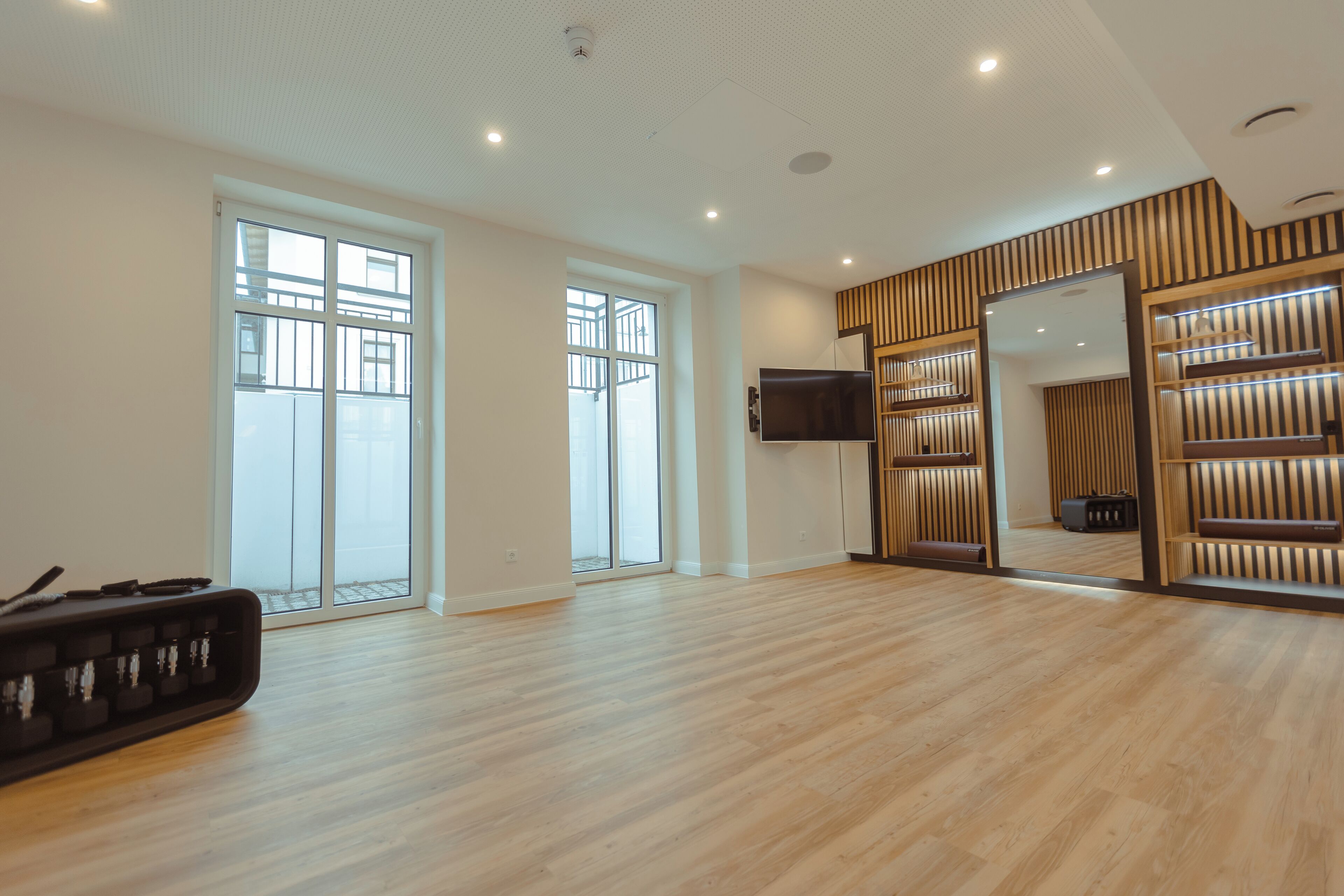 A spacious room with wooden flooring, large windows, a flat-screen TV, and built-in wooden shelving.