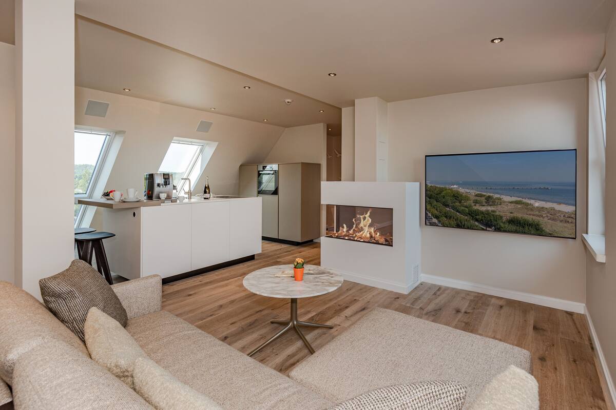 Ein modernes Wohnzimmer mit einer Couch, einem kleinen runden Tisch und einem Kamin. Im Hintergrund ist eine Küche mit einem großen Fenster und einem Kühlschrank zu sehen.