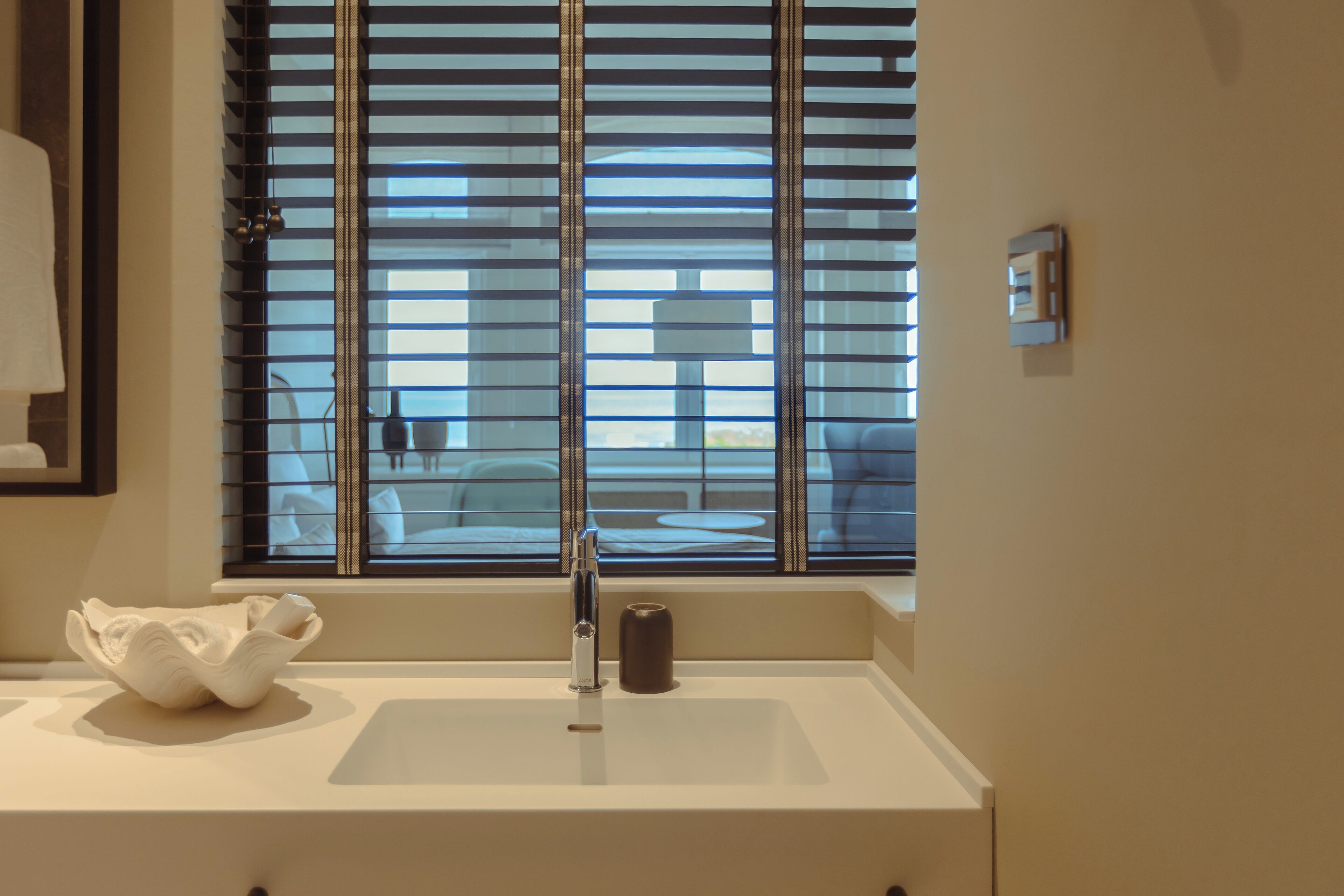 Un baño con un lavamanos blanco, un grifo moderno y una ventana con persianas horizontales.