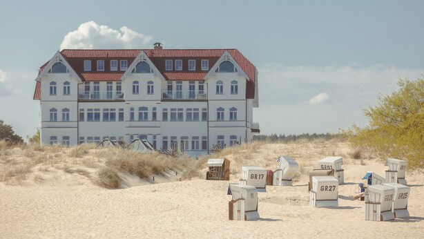 A large, multi-story building with a red-tiled roof, situated on a sandy beach with dunes in the foreground. - Strandhotel Ostende (Heringsdorf)