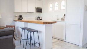 Una cocina moderna con armarios blancos, encimera de madera y microondas integrado. Hay dos taburetes de bar y una mesita con lámpara.