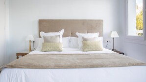 Una cama bien hecha con un cabecero color crema, dos mesitas de noche con lámparas, y una ventana con vistas a zonas verdes.
